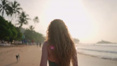 Deniz kenarında huzurlu bir sabah yürüyüşü, sağlıklı bir yaşam tarzı, saç bakımı. Kıvırcık saçlı bir kadın güneş doğarken sahilde yürür, okyanus esintisini hisseder. Yalnız başına gevşer, tropik ada kaçamağı, doğa bağlantısı.