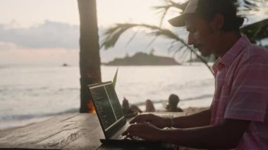 Dizüstü bilgisayarda serbest çalışan kodlar, etrafta palmiye ağaçları, arka planda dalgalar. Gün batımında deniz kenarında Beach Cafe 'de web geliştirici programları. Adam okyanus manzaralı, ücra, manzaralı bir ofiste çalışıyor. Yavaşla