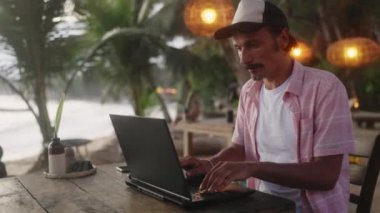 Erkek geliştirici senaryolar, palmiye ağaçları yakınında yazılımlar, okyanus manzarası. Gün batımında plaj kafede dizüstü bilgisayarda şifreler. Serbest programcı deniz yoluyla çalışıyor. Uzak iş yaşam tarzı, dijital göçebe.