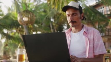 Freelancer deniz kenarında, palmiye ağaçlarında çalışıyor. Tropikal plaj barında dizüstü bilgisayarda şapkalı sıradan bir adam, bir bardak bira bir yana. Dijital göçebe çalışmaktan zevk alır, gün batımı ışık filtreleri yaprakların arasından geçer.