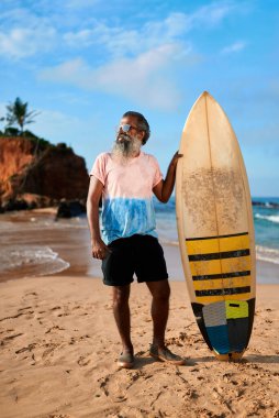 Sörf tahtası olan son sınıf öğrencisi kumlu sahilde duruyor. Yaşlı sörfçüler okyanus kenarında güneşli bir günün tadını çıkarırlar. Yaşlı yetişkinler için aktif yaşam tarzı. Tatil ortamında deniz dalgaları mavi gökyüzü.
