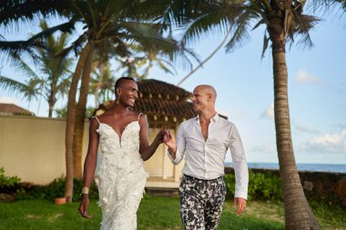 Beyaz elbiseli siyah kişi tropik düğünde takım elbiseli LGBTQ ortağıyla el ele tutuşuyor. Palmiye ağaçlarının altında gülümseyen bir çift. Ayrıcalıklı evlilik, modern aşk çeşitliliği neşe.