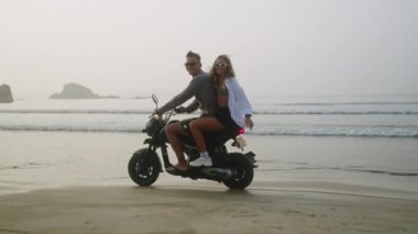 Gün doğumunda deniz kenarında motosiklet süren mutlu çift. Okyanus kıyısında motosiklet süren bir kadın. Bikinili, tişörtlü, güneş gözlüklü güzel bir bayan kameraya bakıp gülüyor, gülümsüyor. Rüzgâr saçlarını savuruyor.