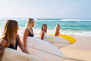 Bir grup sörfçü kadın kumsalda yürüyor. Mayo giymiş gülümseyen arkadaşlar sörf gezisine hazır. Gün be gün, okyanus dalgaları. Kadın sörf macerası seyahati.