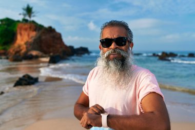 Kıdemli Hintli, kumsaldaki gri sakallı güneş gözlüklü adam akıllı saat kullanıyor. Yaşlı, kablosuz kulaklıkla takip cihazını kontrol ediyor. Tarz sahibi büyükbaba teknolojiden hoşlanır. Yaşlılar için giyilebilir moda araçları.