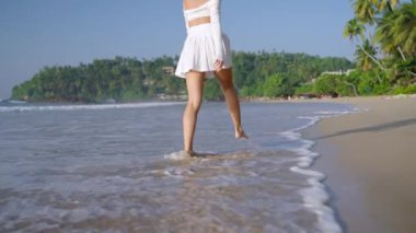 Güzel genç bir kadın kumlu okyanus kıyısında koşuyor. Tropikal adada deniz sörfünde çıplak ayakla yürüyen çekici bir model. İnce kız yaz tatilinde kıyı şeridi dalgalarında eğleniyor..