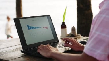 Erkek serbest çalışan, deniz kenarında çalışıyor. Diyagramları gözden geçiriyor. Pro, Sunset Beach Cafe 'deki bilgisayardaki veri tablolarını analiz ediyor. Okyanus manzaralı sıradan bir iş ortamı. Dijital göçebe yaşam tarzı. Yavaş çekim.