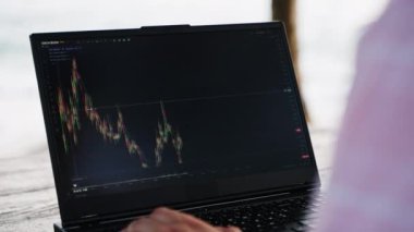 Yetenekli erkek okyanus arkaplanı ve sakin bir akşam atmosferiyle dijital varlık ticareti yapıyor. Kripto tüccarı sahildeki kafede, günbatımında dizüstü bilgisayardaki borsa grafiklerini analiz ediyor. Finansal büyümeye odaklanmış.