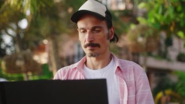 Uzaktan kumandalı iş için günlük kıyafet giyen konsantre bir adam. Palmiye ağaçlarının altında güneşli yapraklarla kaplı. Freelancer, tropik plaj kafesinde dizüstü bilgisayarda çalışıyor. Üretken çevre, doğada rahat çalışma ortamı.