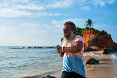 Yaşlı sakallı adam deniz kıyısında akıllı saatle koşar. Sunbae fitness takip cihazı ve kablosuz kulaklık kullanıyor. Sağlıklı yaşam tarzı son sınıfların refahını izler, sahildeki sağlığı izler modern teknolojiyi yönetir..