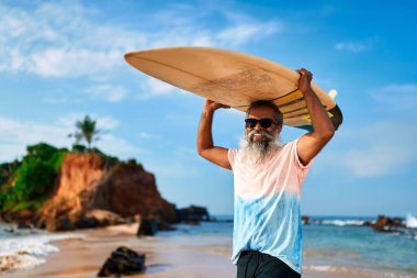 Tropik sahilde sörf tahtası olan yaşlı bir adam. Son sınıf erkek sörfçü güneş gözlüğü takarak gülümsüyor. Aktif yaşam tarzı, okyanus macerası. Sağlıklı yaşlanma, doğanın keyfi. Değişik bir enerji..