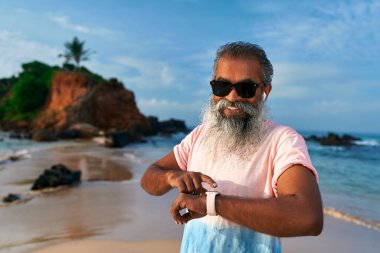 Uzun sakallı yaşlı Hintli adam kumlu sahilde akıllı saat kullanıyor, kulaklıkları kablosuz, gülümsüyor. Sunbae fitness ve sağlık kontrollerini araştırıyor. Modern alet izleri kan şekerini ölçer..