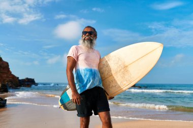 Kumsalda sörf tahtası olan yaşlı bir etnik adam. Aktif yaşam tarzı, okyanus dalgalarında sörf yapmaya hazır. Yaşlı sörfçüler güneşli seyahat yerlerinden hoşlanırlar. Macera turizmine ilham veriyor sağlıklı son sınıf aktiviteleri.