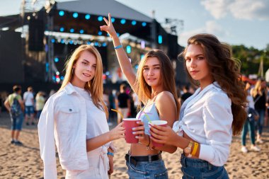 Mutlu arkadaşlar yaz festivali havasının tadını çıkarıyorlar. Arka planda canlı sahnesi olan sıradan bir plaj partisi. Gün batımında kumlu sahilde kokteyllerle kadeh kaldıran bir grup genç kadın açık hava müzik etkinliği sırasında kutlama yapıyorlar..