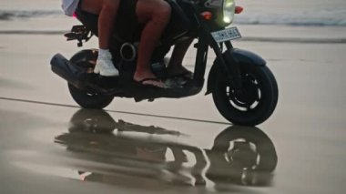 Seyahat eden çiftler gün doğumunda deniz sahilinde motosiklet sürerler. Bisiklet tekerlerinin yansımasından kaynaklanan, insanlara ait bir iz. Turistler okyanus kıyısında kiralık motosiklet kullanıyorlar. Kadın erkeği kucaklıyor..