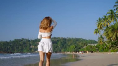 Mutlu bayan okyanus kıyısında neşe içinde koşuyor. Güzel bir kadın gülüyor, deniz kenarında yaz tatilinin tadını çıkarıyor. Gülen manken tropikal kumsalda, palmiye ağaçlarında eğleniyor. Beni takip edin. Yavaş çekim.