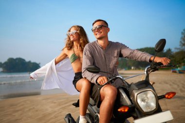 Gülümseyen genç çift tropik plaj boyunca motosiklet sürüyor. Kadın kollarını özgürce açar, erkek scooter kullanır. Macera tatili, deniz melteminin tadını çıkar. Yolculuk için mutlu çiftler.