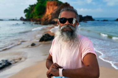 Kıdemli Güney Asyalı adam güneşli sahilde gülümsüyor. Akıllı saat ve kulaklık takıyor. Modern teknolojiyle sakallı, hayat tarzına uygun. Yaşlı yetişkinler yeniliklere kucak açar. Kalpleri genç kalır..
