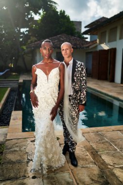 Beyaz elbiseli siyah gelin ve kalın takım elbiseli LGBTQ damat ile villa havuzunda tropik bir düğün. Özel bir aşk kutlaması. Eşsiz olay temaları ya da ilerici düğün promosyonları için mükemmel.