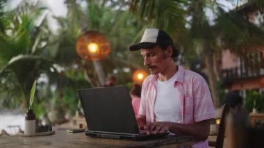 Erkek uzaktan kumandalı işçi projeyle meşgul, okyanus dalgaları, tropikal sahne arkaplanı. Sunset Beach Bar 'da dizüstü bilgisayara odaklanmış serbest çalışan tipler. Sahil manzaralı dijital göçebe yaşam tarzı. Yavaşla