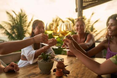 Friends toast wine glasses at sunny outdoor restaurant. Diverse women celebrate bachelorette party trip. Relaxed vibes and fun chat. Stylish setting with palm, tree view. clipart