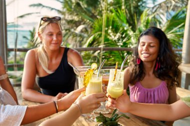 Çeşitli kadın arkadaşlar, deniz kenarındaki restoran masasında, tropikal brunch 'ta içkiyle tezahürat yaparlar. Sosyal zamanın tadını çıkar, gülümseyerek kutla, lezzetli kokteyllerle. Deniz manzaralı, keyifli, keyifli bir deniz yemeği..