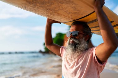 Sörf tahtası olan yaşlı bir etnik adam kumsalda duruyor. Olgun erkek sörfçü, sörf oturumuna hazırlanırken güneşten ve okyanus esintisinden zevk alır. Aktif yaşam tarzı, sağlıklı yaşlanma. Macera yolculuğu titreşimleri.