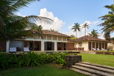 Palmiye ağaçları, taş basamakları ve yemyeşil bahçesi olan klasik tropik bir villa. Beyaz bulutlar güneşli gökyüzü. Çimlerde boş güneşlikler. Tatil inzivasına çekildi. Sessiz cennet ortamı. Mimarlık doğayı karıştırır..