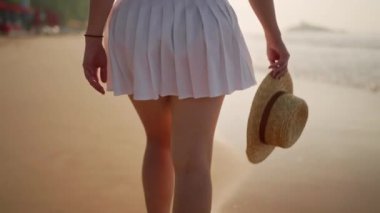 Beyaz etekli kadın deniz kenarında gezmekten hoşlanır, dalgalar yavaşça kıyıya vurur. Gün batımında kumsalda yürüyen bir kadın hasır şapka tutuyor. Tatil gezisi, sakin deniz manzarası.