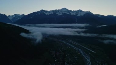 Gün doğmadan önce Mestia Havaalanı 'nın üstündeki dağ vadisinde hava manzarası. Karla kaplı tepeler, alçak bulutlar ve sabah ışığında nehir görünen küçük bir kasaba. Sakin dağ manzarası. İHA atışı