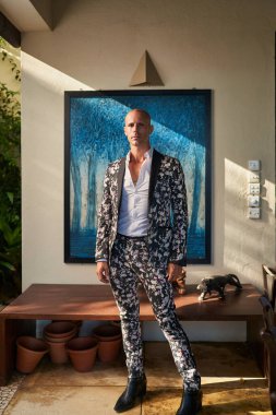 Kalın çiçekli takım elbiseli adam villa avlusunda güvenle duruyor. Güneş ışığı duvar sanatına gölge düşürür. Zarif eşcinsel damat kıyafeti. Moda anlayışı. Potansiyel düğün, moda, yaşam tarzı etkinliği.