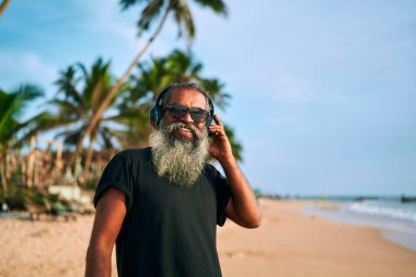 Gri sakallı ve güneş gözlüklü kıdemli stil sahibi adam tropik plajda müzik dinlemekten hoşlanıyor. Aktif yaşlılar kulaklıkları dinliyor, teknolojiyi kucaklıyor. Modern büyükbaba titreşimleri. Okyanus palmiyelerinin yanında rahatla. Canlı enerji..