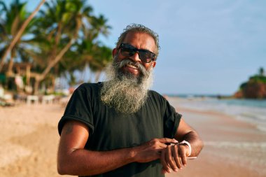 Güneş gözlüklü şık gri sakallı son sınıf öğrencisi tropik plajlarda akıllı saat kullanıyor. Modern teknoloji meraklısı büyükbaba aktif yaşam tarzını ve güneşli günü seviyor. Yaşlı adam kucaklıyor, dijital zindelik.