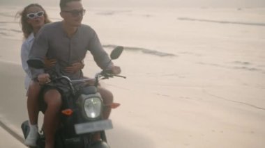 Şık bir çift gün doğumunda deniz sahilinde motosiklet sürüyor. Bisikletten insana aktarma kurulumu. Turistler okyanus kıyısında kiralık motosiklet kullanıyorlar. Kadınlar erkeklere sarılır, motosiklette kollarını açar..