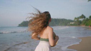 Yalnız gezgin deniz kenarından zevk alır, güneş tarafından öpülen saçların canlılığı özgürlük yayar. Beyaz elbiseli kadın tropik plajda oynaşıyor, gür bukleler enerjik bir şekilde zıplıyor. Şapka elinde, kameraya dön, tamamen rahatla..