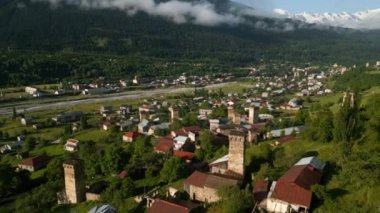 Gün doğumunda bir dağ vadisindeki Svan kulelerinin hava manzarası. Geleneksel taş kuleler, yemyeşil bir manzarada, evler arasında yer alır. Arka planda karla kaplı dağlar var. İHA atışı