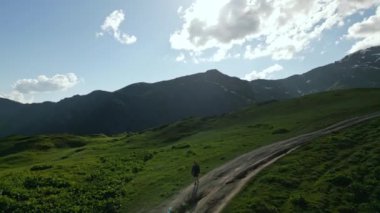 Yüksek dağlardaki alp çayırlarında yürüyüş yapan bir adamın havadan görünüşü. Solo yürüyüşçüler, yeşil çimlerle çevrili patikada yürürler. Arka planda karlı tepeler, berrak gökyüzü. İHA atışı