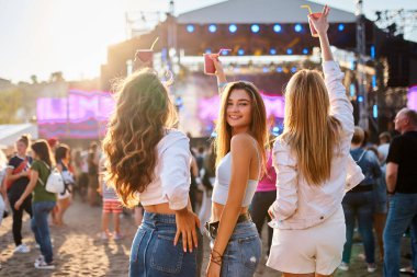 Güneşli sahil etkinliğini kutlamak için gün ışığı kalabalığı toplandı. Bir grup kız, kumlu sahilde yaz müzik festivalinin tadını çıkarıyorlar. Renkli içkiler içiyorlar, gülümsüyorlar ve arka planda dans ediyorlar..