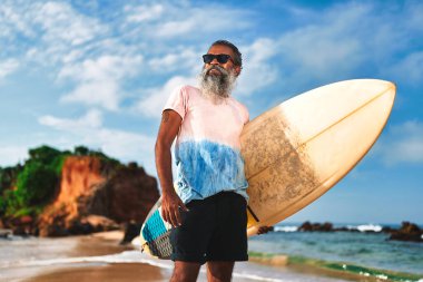 Sörf tahtası olan yaşlı siyah adam güneşli sahilde duruyor. Güneş gözlüklü son sınıf öğrencisi okyanus dalgalarında sörf yapmaktan hoşlanıyor. Yetişkinler için aktif yaşam tarzı macerası.