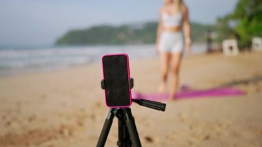 Huzurlu denizin arka planı, hayranlar sağlık ve denge için sanal sınıfı takip ediyor. Spor hocası tripodda akıllı telefon kullanarak canlı plaj yoga seansını yönetiyor. Şafakta canlı sağlık egzersizi.