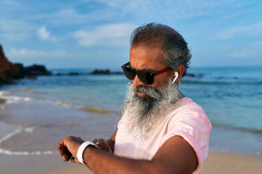 Yaşlı sakallı adam sahilde akıllı saat kullanır. Kablosuz kulaklıklı dedem modern teknolojinin keyfini çıkarıyor. Güneş gözlüklü olgun bir adam takılabilir aletleri kontrol eder. Sağlıklı bir yaşam tarzı. Okyanus arkaplanı.