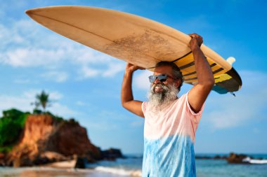 Güneşli okyanus sahilinde sörf tahtası olan yaşlı siyahi bir adam. Aktif yaşlı erkek dalgalarda sörf yapar, kıyı yaşamından zevk alır. Fit yaşlı sportif maceraya atılır sağlıklı emeklilik.