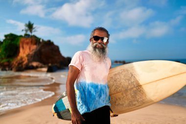 Kumsalda sörf tahtası olan yaşlı bir etnik adam. Olgun sörfçü aktif yaşam tarzını sever, işten sonra dinlenir. Emeklilik, eğlence, spor, özgürlük, okyanus manzarası. Sağlıklı yaşlanma, altın yıllar kıyı yaşamı.