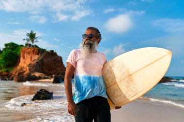 Gri sakallı yaşlı adam okyanus kıyısında sörf tahtası tutuyor. Tropikal plajda aktif yaşam tarzı. Yaşlı sörfçü güneşli havada kıyı macerasından hoşlanır. Yaşlılar için seyahat turu, sörf içeriyor.