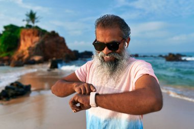 Güneş gözlüklü gri sakallı yaşlı adam sahilde akıllı saat kullanıyor. Kulaklık takan olgun bir erkek sağlık kontrolü yapıyor. Kablosuz teknik kullanım, refah için yaşlılar.