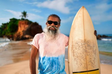 Sunny Beach 'te sörf tahtası olan yaşlı sakallı bir adam. Yaşlı sörfçü okyanus kenarında aktif yaşam tarzını seviyor. Güneş gözlüklü yaşlı erkek, deniz maceraları ve zindeliği kucaklıyor. Seyahat eğlencesi..
