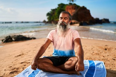 Kumsalda yaşlı adam yogası Lotus Pozu. Meditasyon, gün batımında kayalık adanın yanındaki okyanus kıyısında farkındalık. Sabah sağlığı, rahatlama. Sunbae, deniz kıyısındaki huzurun tadını çıkarıyor..
