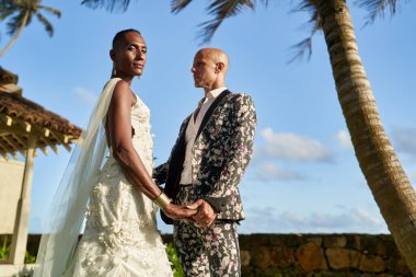 Düğünde tropikal villada çift el ele tutuşuyor. Çiçek takım elbiseli beyaz elbiseli gelin. LGBTQ teması. Cennetin güneşli yerinde seremoni.