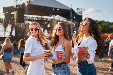 Renkli içkiler içip, arka planda dans ederek. Güneş gözlüklü bir grup mutlu genç kadın kumsalda yaz müzik festivalinin tadını çıkarıyor. Günlük kıyafetler, moda aksesuarlar, hareketli kalabalık..