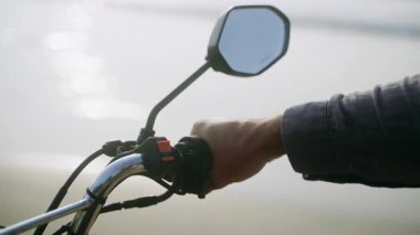 Bisikletçi, deniz kenarındaki bisiklet gidonuna elini koydu. Sahilde motosiklet kullanan biri var. Motosiklet konsolu, dikiz aynaları. Okyanus kıyısında scooter 'da oturan adam.